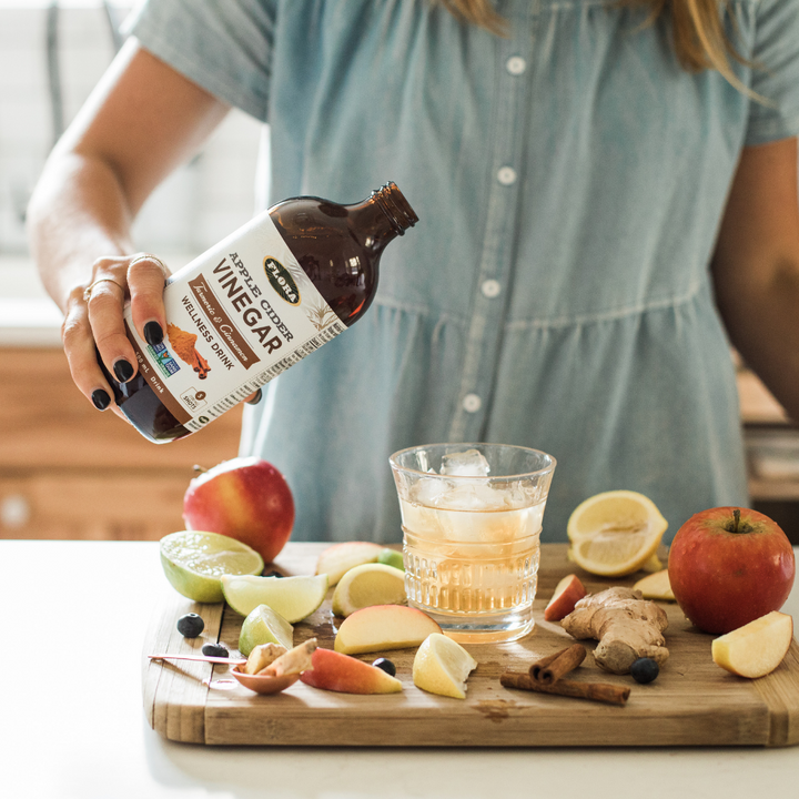 Flora organic, non-gmo apple cider vinegar with cinnamon and turmeric poured into glass of water surrounded by fresh fruit and herbs: apples, ginger, lemons, cinnamon, limes, and elderberries